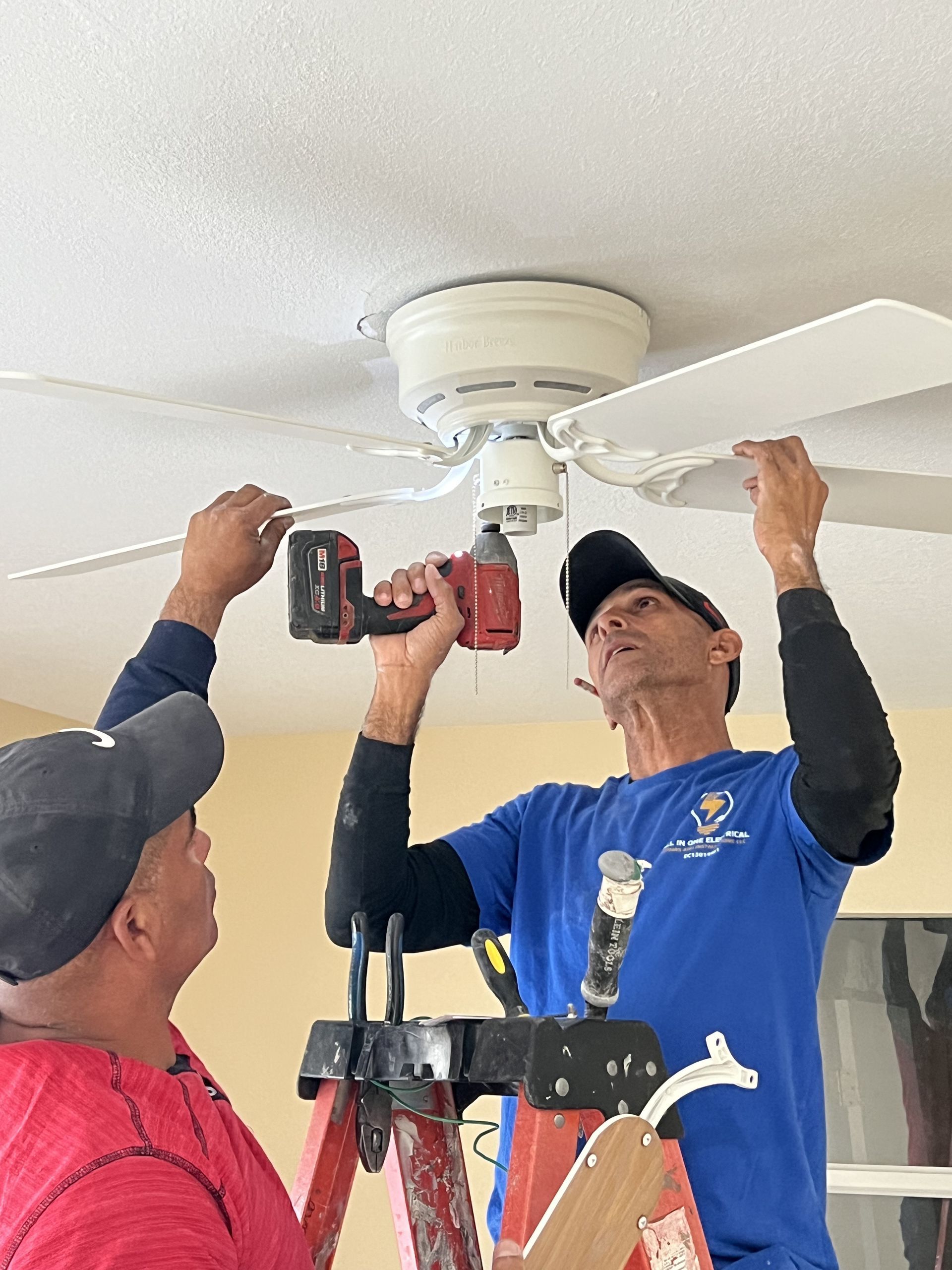 Two men on a ladder installing a white ceiling fan. One uses a drill; both are wearing hats and focused.
