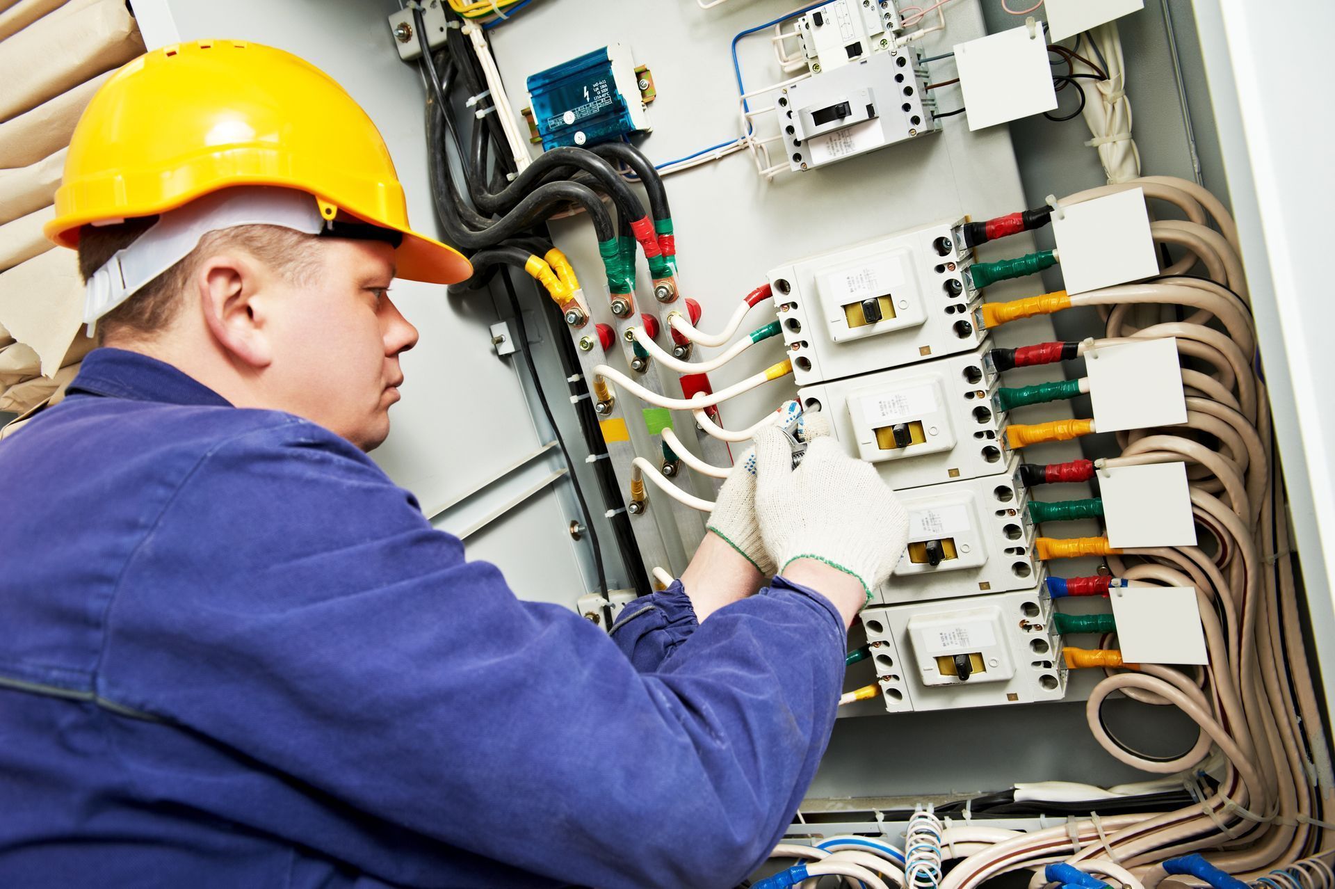 Electrician in blue uniform and yellow hard hat working on electrical panel.