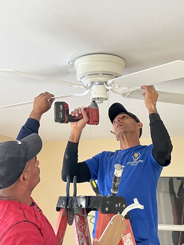 Two men on a ladder installing a white ceiling fan, one using a drill.