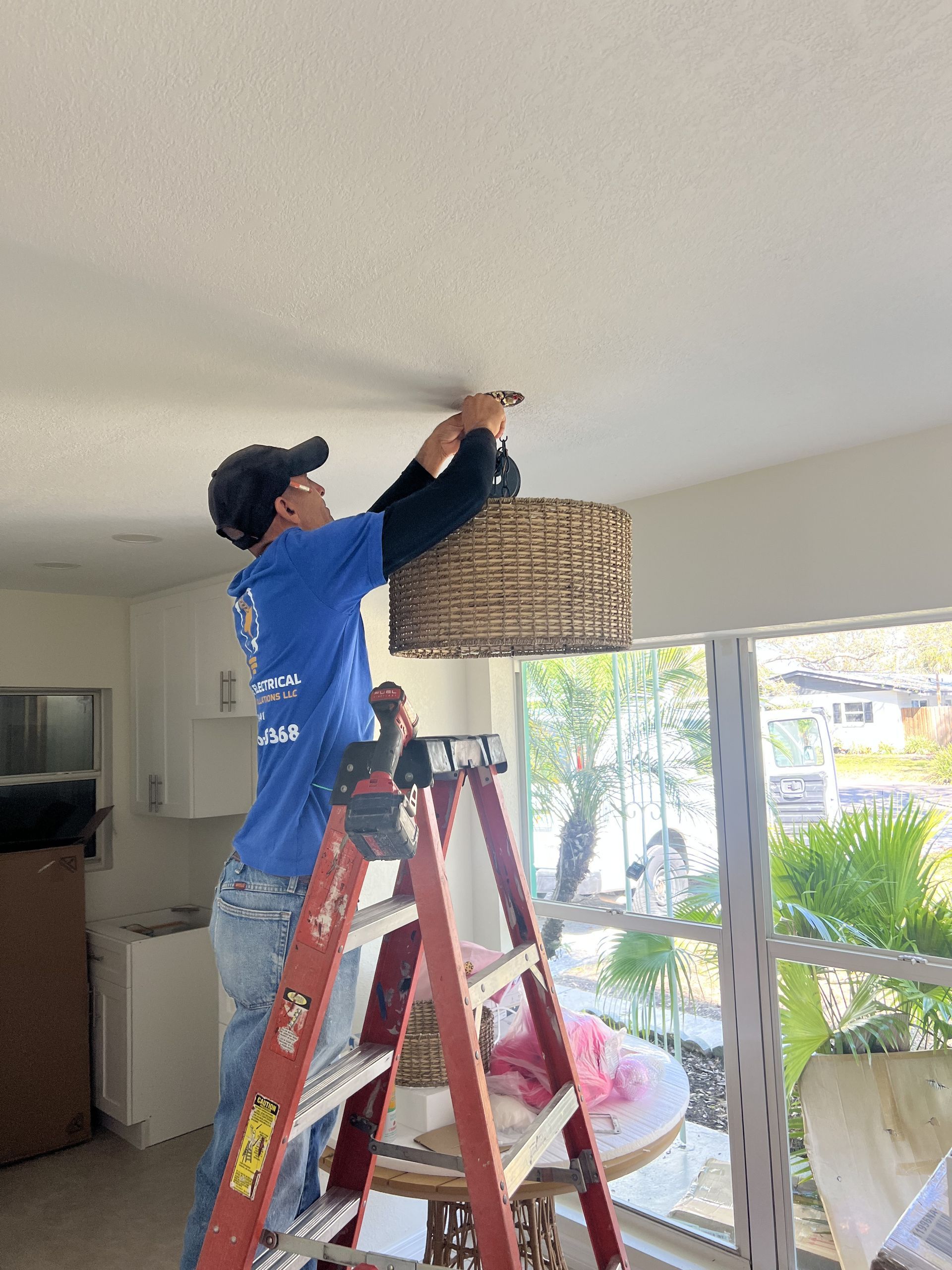 Man on a ladder installing a woven lampshade on a white ceiling. Kitchen and trees visible.