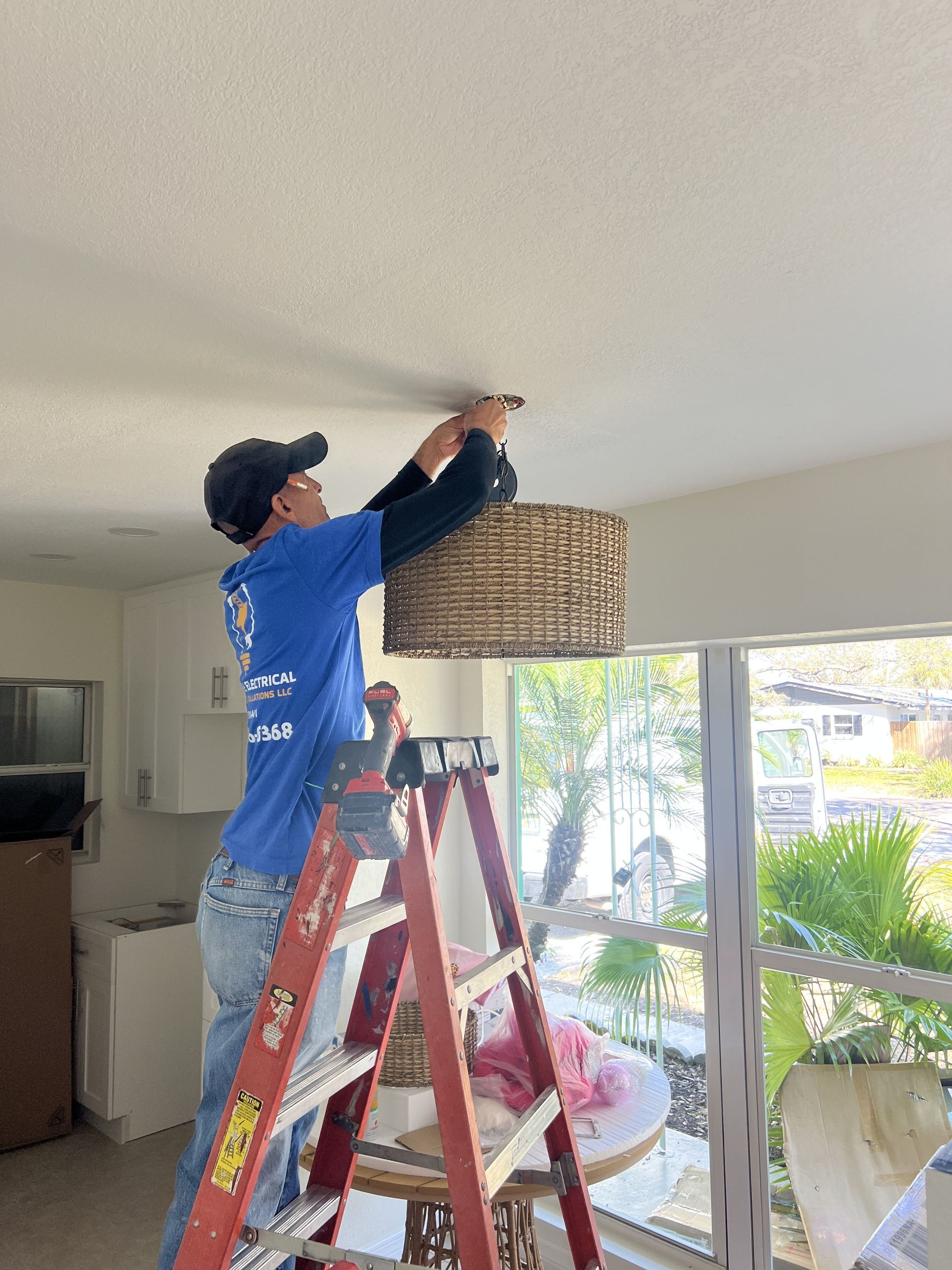 Man in blue shirt on ladder installing a wicker lampshade on the ceiling indoors.