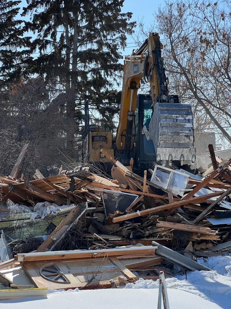 A yellow excavator demolishes a wood-framed house into a pile of debris on a snow-covered lot.