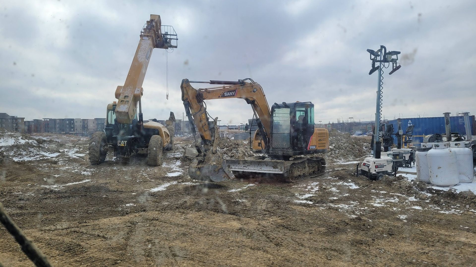 A construction site with a yellow excavator and a crane parked on a dirt lot under an overcast sky.