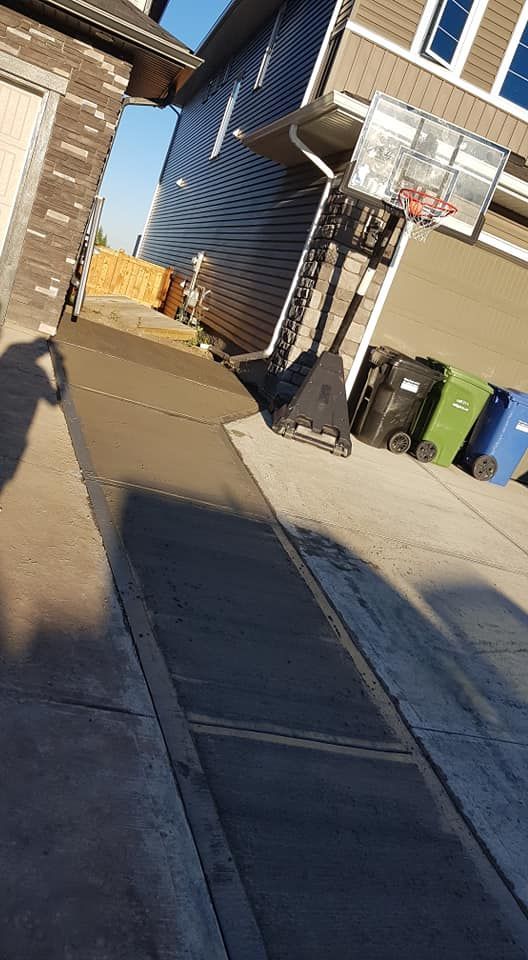 A concrete walkway leading to a wooden ramp between two houses, with a basketball hoop and garbage bins nearby.