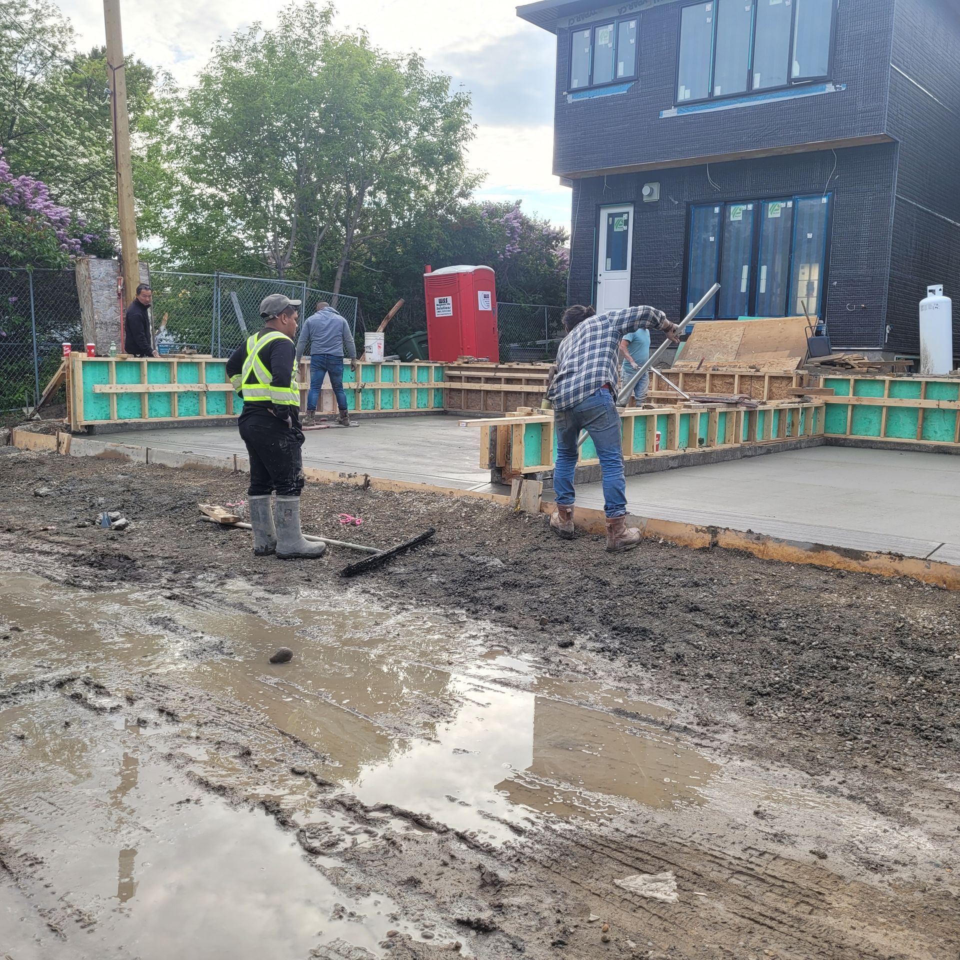 Construction workers in safety gear finish a concrete foundation at a residential site near a muddy, water-filled area.