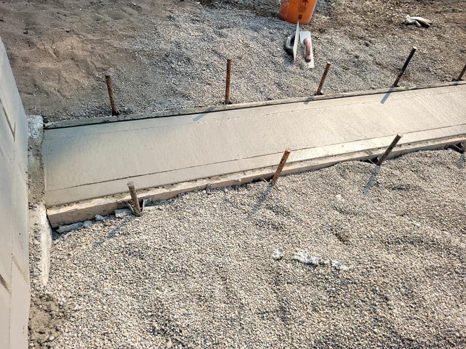Freshly poured concrete walkway section under construction, framed by wooden boards and metal stakes in a gravel area.