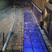 A construction site showing rebar grid laid over blue vapor barrier sheets, prepared for pouring a concrete walkway.