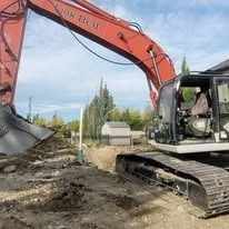 An orange Link-Belt excavator sits on a dirt lot, its arm raised over an excavated trench in a residential area.