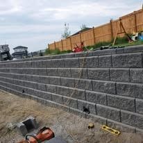 A newly constructed gray concrete block retaining wall stands outdoors, with construction tools visible on the ground.