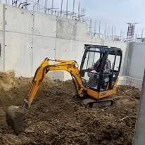 A yellow compact excavator digging in a dirt pit at a construction site with concrete walls in the background.