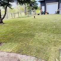 A person mows a green lawn near a large building with a closed garage door on a sunny day.