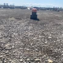 An orange piece of construction machinery sits on a wide, rocky, uneven dirt field under a bright, clear sky.