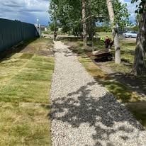 A gravel path stretches between a green fence and trees, with fresh sod being laid on the left.
