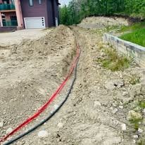 A long, red conduit and a black utility cable lay in a dirt trench leading toward a house.