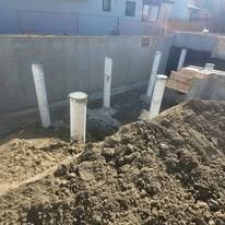 Vertical concrete piers rise from a muddy construction site in front of a partially finished grey concrete foundation.