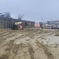 A reach forklift carries a load in front of a tall, gridded concrete retaining wall at a muddy construction site.