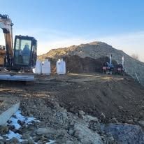 An excavator parked on a dirt construction site near white storage tanks and equipment against a large, hilly mound.
