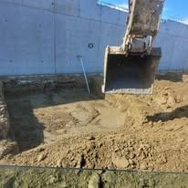An excavator bucket hangs over an excavated dirt construction site in front of a concrete wall.