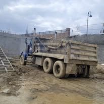 A blue dump truck is parked on a muddy construction site next to a concrete wall and a metal ladder.