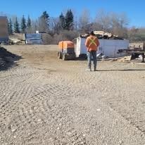 A worker in a high-visibility orange jacket stands on a gravel construction site near a trailer and equipment.