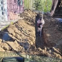 A person in a light-colored coat works in a deep dirt trench next to a house, holding a long measuring pole.