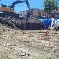 A yellow excavator sits atop a large pile of dirt at an active construction site, beside a blue tarp and white pipes.