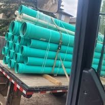 A flatbed truck loaded with a large stack of bright green PVC sewer pipes secured with straps.