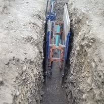A metal trench box secured within a narrow, excavated dirt trench, with a utility pipe visible at the bottom.