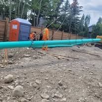 Workers in safety gear guide a long turquoise pipe suspended by a crane on a rocky construction site with a portable toilet.