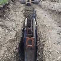 A vertical view of a narrow excavation trench containing metal shoring panels and struts, with an excavator parked above.