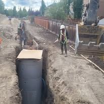 A construction worker in a hard hat and safety vest stands near a deep trench with a large vertical pipe segment.