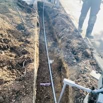 A narrow trench in the ground containing a conduit pipe, with a person standing nearby on the edge.