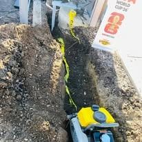 A small yellow and gray gas-powered trenching machine sits in an excavated dirt trench lined with yellow caution tape.