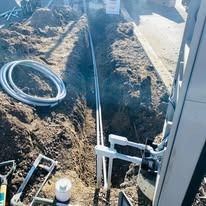 A trench with two white conduit pipes running through it, next to a coil of pipe and electrical equipment.