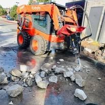 An orange Kubota skid-steer loader uses a hydraulic breaker attachment to demolish concrete pavement outdoors.