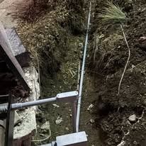 Gray electrical conduit runs through a narrow dirt trench alongside a stone wall foundation.