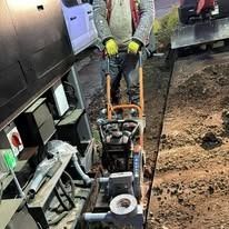 A person operates a walk-behind compactor to press down dirt in a trench next to a utility vehicle.