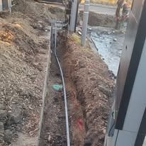 A deep construction trench with a conduit pipe running along the bottom, viewed from above near a utility pole.