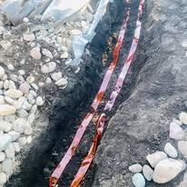 A narrow, dark trench in the ground contains two parallel strips of metallic orange warning tape buried in the soil.