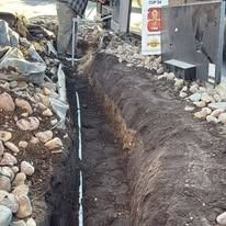 A person in work clothes stands by a narrow, freshly dug trench containing a white pipe, bordered by large landscaping rocks.