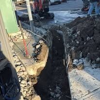 A narrow trench dug into the ground at a construction site, with exposed dirt, rocky debris, and equipment nearby.