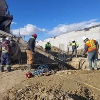 Construction workers in high-visibility gear guide a concrete chute at a building site under a bright blue sky.