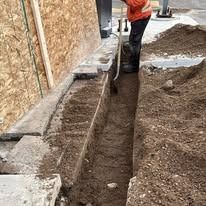 A person in high-visibility orange clothing using a shovel to dig a narrow trench alongside a plywood wall and dirt pile.