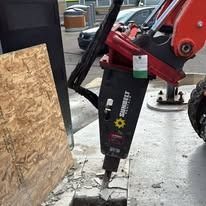 A Sunbelt Rentals hydraulic hammer attachment on an excavator breaks up a concrete sidewalk near a plywood barrier.