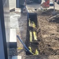 A narrow trench in the ground containing yellow caution tape, leading toward a metal utility pedestal at a construction site.