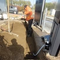A person in an orange work jacket digs a trench in the dirt next to a metal gate controller pillar.