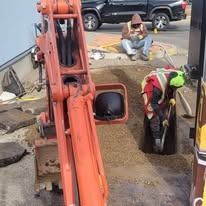 A person in a high-visibility vest digs in a trench next to an orange excavator, while another person sits nearby.