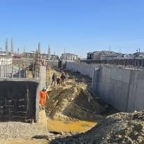 Construction workers on a muddy, dirt-filled site between large concrete foundation walls under a bright blue sky.