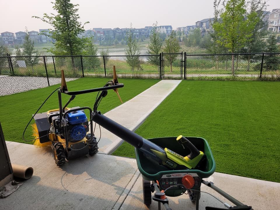 A power broom and a lawn spreader with a leaf blower sit on a concrete patio next to a manicured artificial lawn.