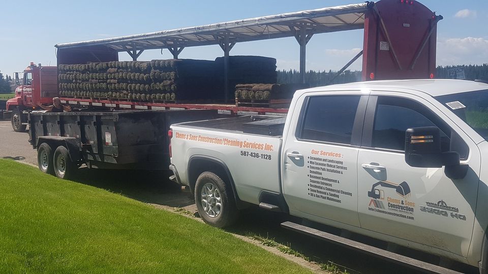 A white pickup truck with company decals parked on grass, towing a small trailer in front of a flatbed truck on a sunny day.
